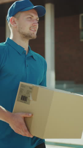 Handsome Delivery Man Holds Cardboard Box Package Walks Through Modern Stylish Business District. Low Angle Slow Motion. Vertical Screen Orientation Video 9:16