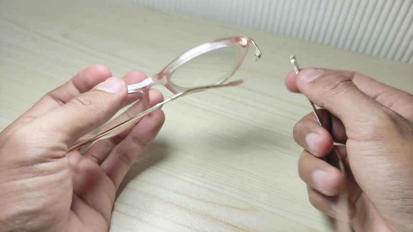 Close-up of hands carefully inspecting a broken right temple (handle) of a pair of eyeglasses over a wooden tabletop