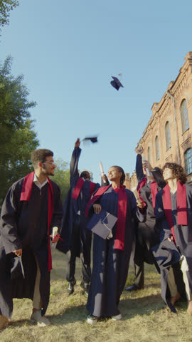 Vertical shot of cheerful group of diverse students throwing their hats up after graduation ceremony outdoors