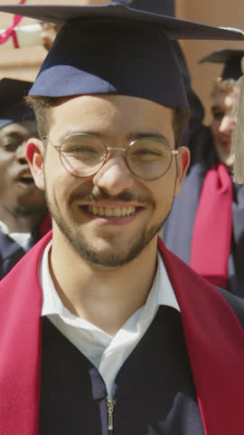 Vertical portrait of positive graduate in hat and gown looking at camera while his collegemates celebrating in background