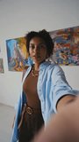Medium vertical selfie shot of young African American woman shooting video on smartphone for social media while visiting contemporary art exhibition in museum, and showing paintings to followers