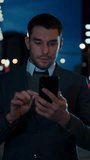 Caucasian Businessman in a Suit is Using a Smartphone on Dark Street in the Evening. Other Office People Walk Past. Video Footage with Vertical Screen Orientation 9:16.