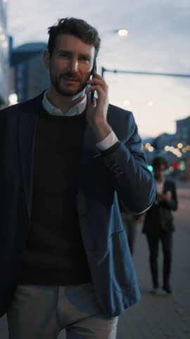 Businessman in a Suit is Talking on a Phone While Walking in the City. He's Having a Pleasant Conversation. Office People Walk By. It's Evening. Video Footage with Vertical Screen Orientation 9:16. Royalty-Free Stock Footage #3462233143