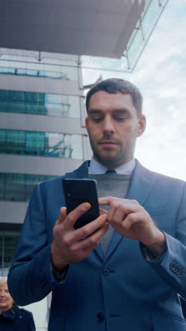 Caucasian Businessman in a Suit is Using a Smartphone on Dark Street in the Evening. Other Office People Walk Past. Video Footage with Vertical Screen Orientation 9:16. Royalty-Free Stock Footage #3462240477