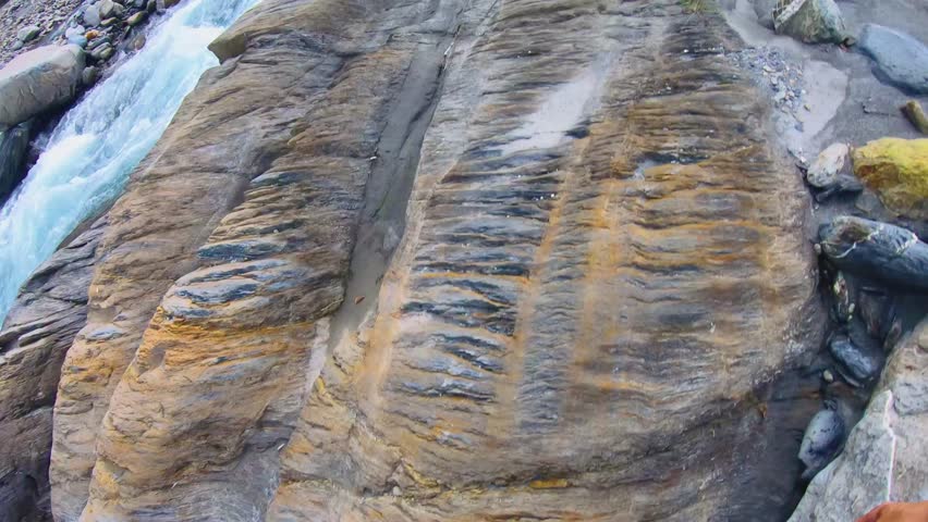slow motion of walking up to brown boulder of rock with pile of gravel near a river water surged rapidly with speed raced downstream with mountains creek valley canyon hills by the riverbed riverside