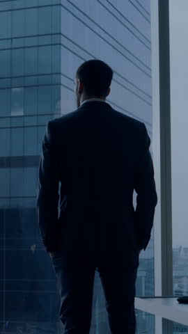 Back View of the Thoughtful Businessman wearing a Suit Standing in His Office, Contemplating Next Big Business Deal, Looking out of the Window. Video Footage with Vertical Screen Orientation 9:16