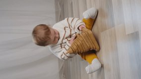 Vertical video of beautiful little male toddler playing with wooden cube sitting on warm floor at home. Curious boy enjoying his toy. Concept of innocence and childhood