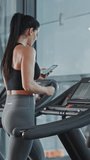 Beautiful Athletic Sports Woman in the Gym Puts on Wireless Headphones, Turns on Podcast or Sport Music Playlist with Smartphone and Starts Running on a Treadmill. Vertical Screen