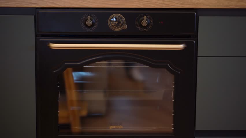 Cesis, Latvia - February 14, 2024 - Close-up of a modern black oven with a glass door and gold handle, set in dark cabinetry with wood countertops, and control knobs with an analog clock. Royalty-Free Stock Footage #3462355435