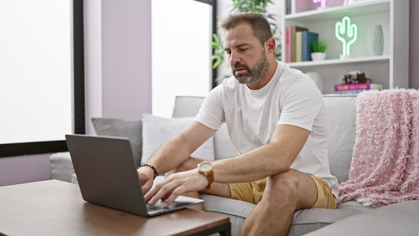 Middle-aged man with grey hair using a laptop on a sofa, then relaxing and covering with a cushion in a modern living room. Royalty-Free Stock Footage #3462357453