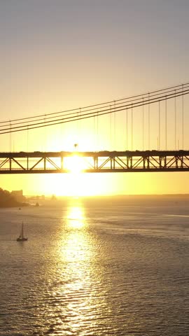 Silhouette of Ponte 25 de Abril Suspension Bridge at Sunset and Sailing Yacht. 25th April Bridge. Portugal. Aerial View. Orbiting. Vertical Video