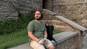 Video 4K. Young adult bearded white man with long hair in shorts sits on wall and drinks water from plastic bottle on old street of Georgian city of Tbilisi. Summer sunny day. Evening sun. Golden hour