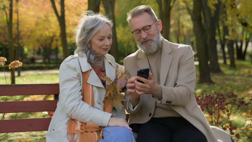 Caucasian retired couple family in love mature woman man sit on bench autumn park using mobile phone gadget together smiling wife husband spouses reading news old age and smartphone in city outdoors