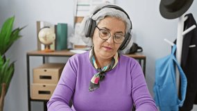 A smiling grey-haired woman in headphones sits in an office, her vibrant scarf contrasting with the professional atmosphere.