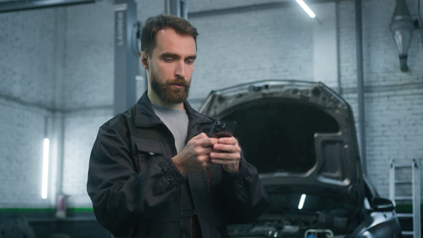 Mechanic man auto mechanical worker using mobile phone gadget in repair car service garage automobile factory pensive thoughtful male guy repairman look at smartphone screen think typing on cellphone Royalty-Free Stock Footage #3462623647