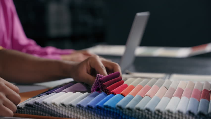 Close up hands fashion designer tailor choose textile samples cloth fabric collection. Cropped shot unknown man choosing material color for design sewing clothes in atelier while woman typing laptop