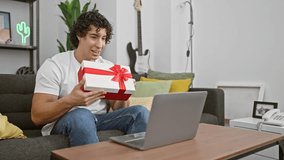 A cheerful young man in a casual setting at home, interacting with a laptop and holding a red gift box.