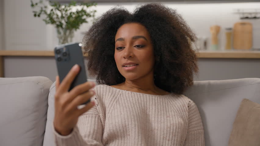 African American woman smiling carefree girl ethnic female lady homeowner on sofa talk video call smartphone chatting distant communication with mobile phone at home apartment positive virtual meeting Royalty-Free Stock Footage #3462657809