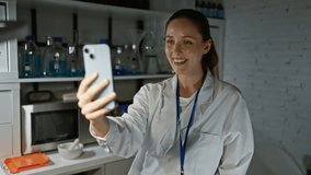 Smiling woman scientist in lab coat making a video call with a smartphone in a laboratory setting