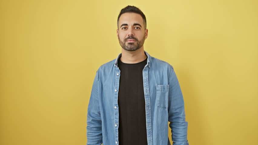 Young hispanic man wearing denim shirt standing hand on mouth telling secret rumor, whispering malicious talk conversation over isolated yellow background