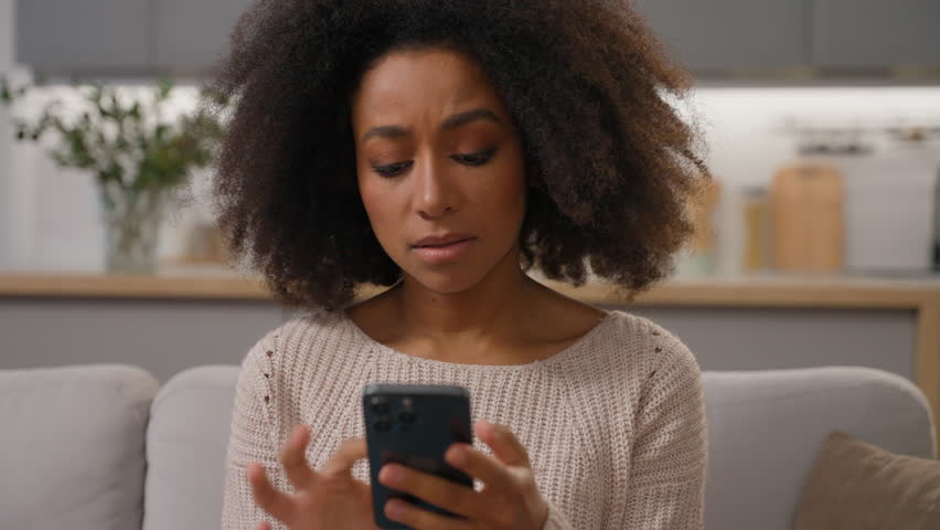 Shocked sad girl African American woman on sofa hold smartphone looking at mobile phone screen worried read bad news failure negative result rejection bank debt cellphone connection problem at home Royalty-Free Stock Footage #3462671287