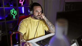 A bearded hispanic man enjoys gaming at night in a neon-lit room wearing headphones and a casual outfit.