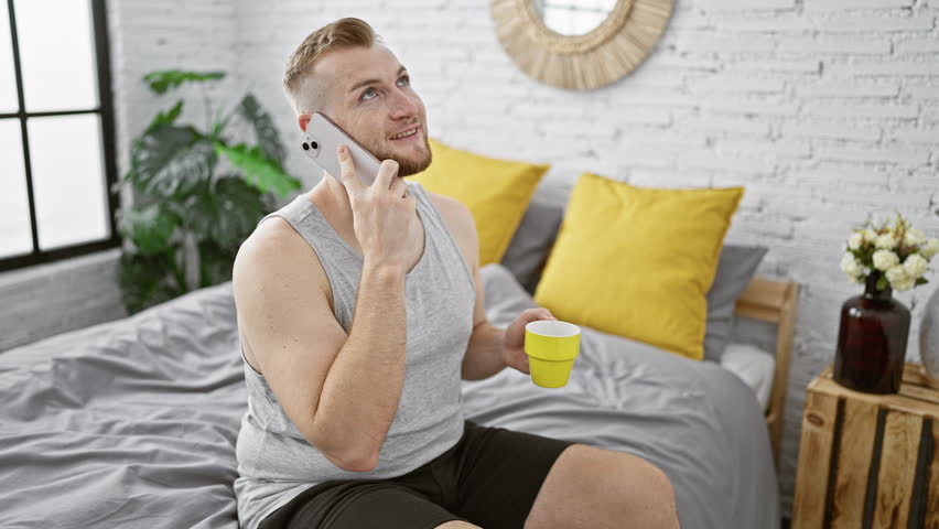 A smiling young man with a beard enjoys a morning phone call while holding a cup in a stylish bedroom.