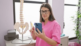 Hispanic woman healthcare professional in pink scrubs using smartphone in clinic with spine model.