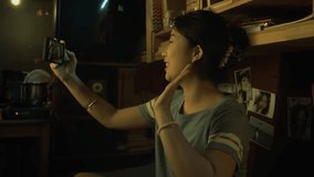 Medium close-up shot of cheerful young Chinese woman sitting on bunk in shabby micro flat, holding out smartphone, waving then talking on video call with girlfriends or relatives