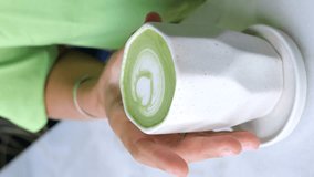 Vertical video woman holding large white mug with aromatic matcha latte. Close-up matcha bright green color standing out against milk foam. Matcha is popular drink that combines taste health benefits.