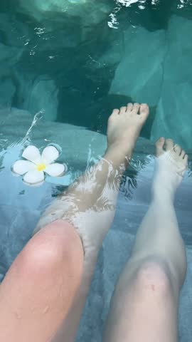 Top view woman bare legs playing in swimming pool or sea or pond surrounded by splash white frangipani flowers floating on the blue water surface