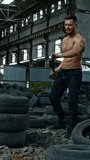 Sportsman with a heavy hammer in old plant. Sporty man trying to break pile of car tires with a metal hammer and smiles to camera. Workout in abandoned place. Vertical video