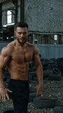 Shirtless bodybuilder in abandoned place. Young athlete with trained muscles posing to camera on used car tires background. Vertical video