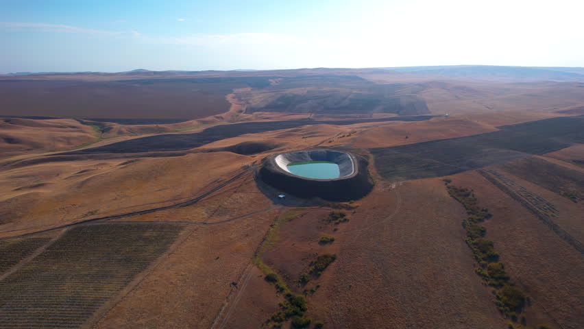 Aerial view of a circular water reservoir in a dry landscape at sunset. A drone captures a circular reservoir amidst dry fields as the sun sets, casting warm hues over the land. Royalty-Free Stock Footage #3463063515
