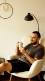 a bearded guy in a gray t-shirt and shorts sits in a chair at home under a floor lamp and drinks tea and chats and laughs. vertical video