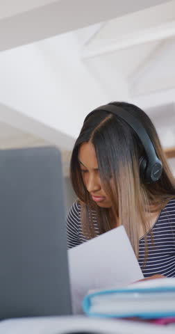 Vertical video of biracial teenager girl wearing headphones and using laptop, slow motion. Spending quality time at home, domestic life and friends concept.