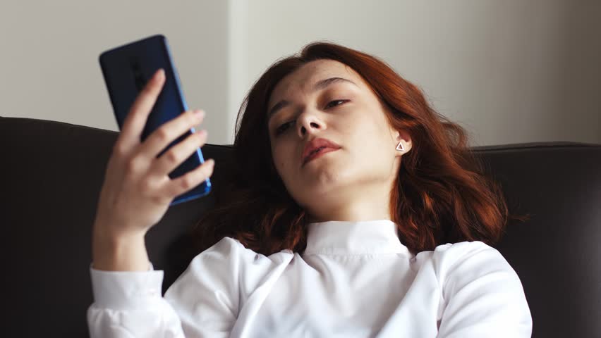 Sad upset dissatisfied boring young woman at home sitting on sofa spending time holding mobile cell phone. Bad mood lifestyle concept