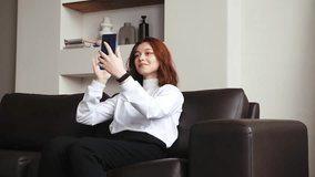 young relaxed woman with sits in comfortable sofa in living room, relaxing at daytime, uses mobile phone for video call and talks smiling and positively. influencer on online webinar