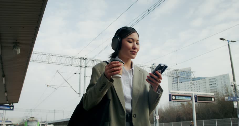 A woman in formal clothes and with headphones on her head uses a mobile phone and drinks coffee from a disposable cup walking along the railway platform