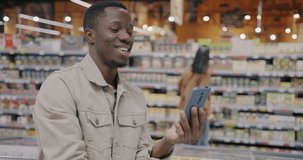 Joyful African American man showing food in supermarket making online video call with smartphone talking. Communication and shopping concept.