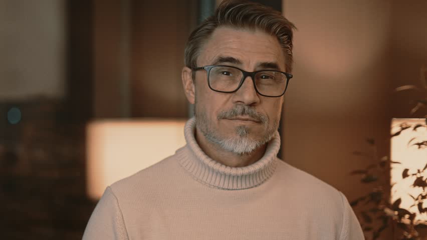 Portrait of confident, elegant middle aged man smiling, standing in living room at home or in hotel apartment. Happy male in glasses and turtleneck pullover. Royalty-Free Stock Footage #3463317913