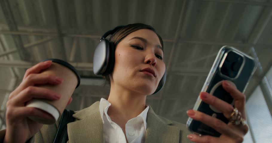A woman in formal clothes and with headphones on her head uses a mobile phone and drinks coffee from a disposable cup walking along the railway elevated passage