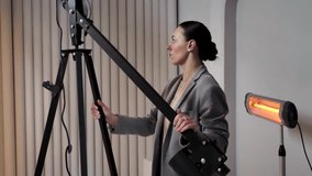 A young woman photographer in a gray suit adjusts professional lighting equipment for an indoor photo shoot. She is seen adjusting the height of a tripod and positioning a halogen lamp. 