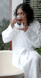 Vertical video of biracial woman sitting on bathtub drinking tea at home, slow motion. Lifestyle, relaxation, self care and domestic life, unaltered.