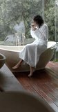 Vertical video of biracial woman sitting on bathtub drinking tea at home, slow motion. Lifestyle, relaxation, self care and domestic life, unaltered.