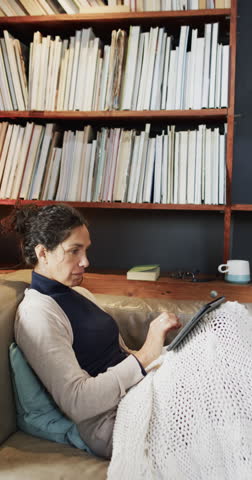 Vertical video of happy biracial woman lying on sofa and using tablet at home, slow motion. Lifestyle, free time, relaxation, communication and domestic lifestyle, unaltered.