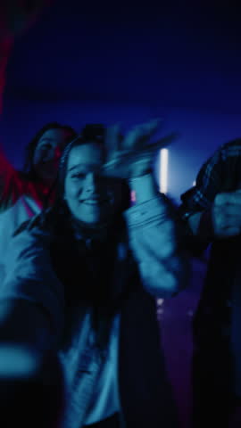 Vertical POV Selfie Portrait of Three Happy and Smiling Stylish Young Adults Waving at the Camera. Two Stylish Woman and a Handsome Man in Futuristic Virtual Neon Garage Studio Environment