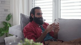Man sitting on sofa, scrolling on smartphone in living room. Video of handsome indian man, phone in hand, smiling.