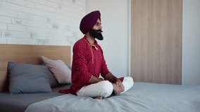Man sitting in bedroom praying, meditating at home in the morning. Video of handsome Indian man with turban.