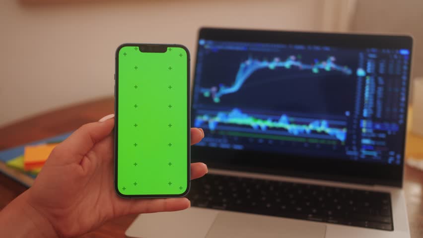 Trader holds smartphone in hands with green screen on chart of movement of cryptocurrency on screen of monitor laptop background. Stock market data statistics using mobile app. Royalty-Free Stock Footage #3463696783